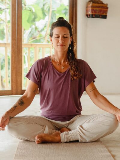 Yogini assisse en méditation sukhasana qui porte un pantalon large confortable écru et un top de yoga violet au décolleté léger resserré aux hanches