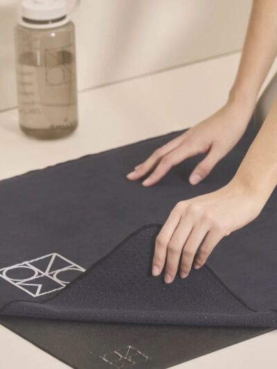 Serviette de yoga noire avec logo Monnchild blanc déposée sur un tapis de yoga noir avec gourde bouteille d'eau