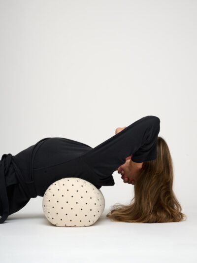 Femme allongée sur le dos qui soutient sa tete dans ses mains avec un bolster de yoga rond écru à pois noirs posé en travers sous ses omoplates